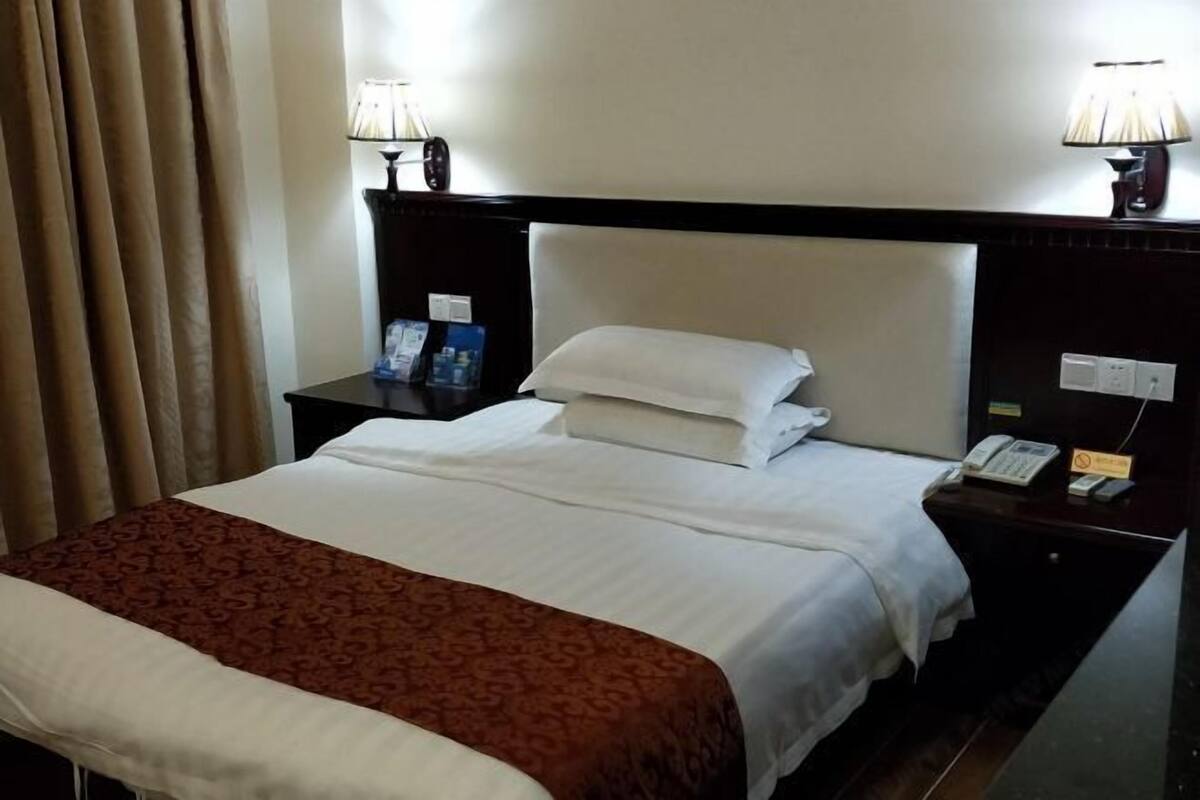 A hotel room with a bed, a nightstand with a lamp, a phone, and a sign.