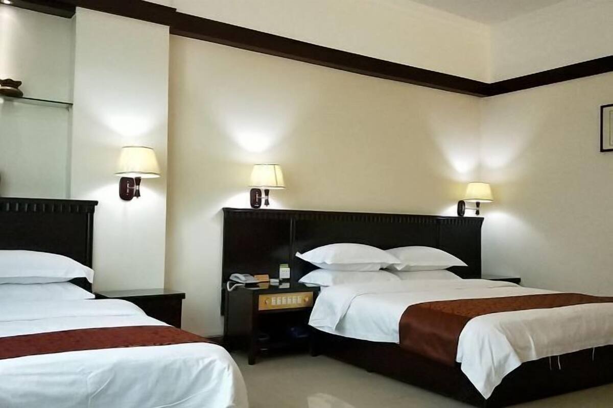 A hotel room with two beds, each with white bedding and a brown throw blanket. There are bedside tables with lamps and a small nightstand with a telephone.