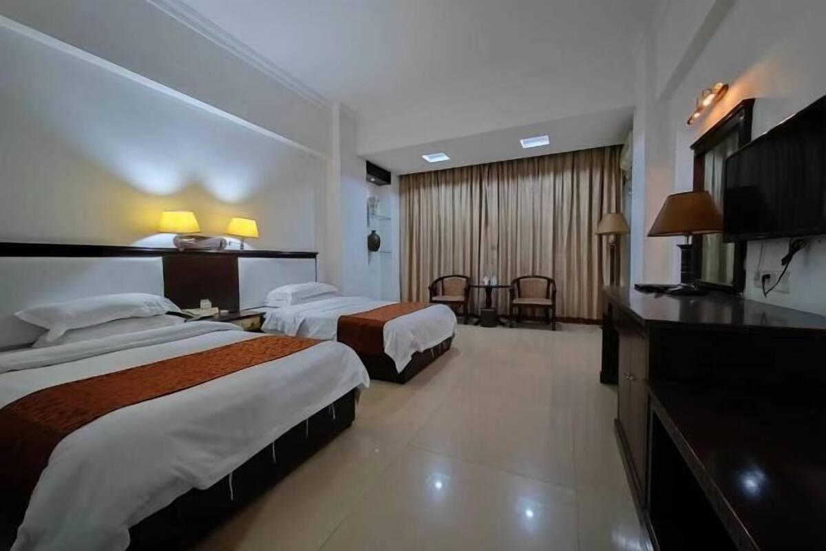 A hotel room with two beds, a desk, a TV, and a seating area with curtains.