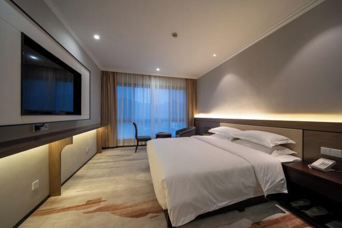 Ein Hotelzimmer mit einem großen Bett, einem an der Wand montierten Fernseher, einem Schreibtisch mit Stuhl und einem Fenster mit Vorhängen.