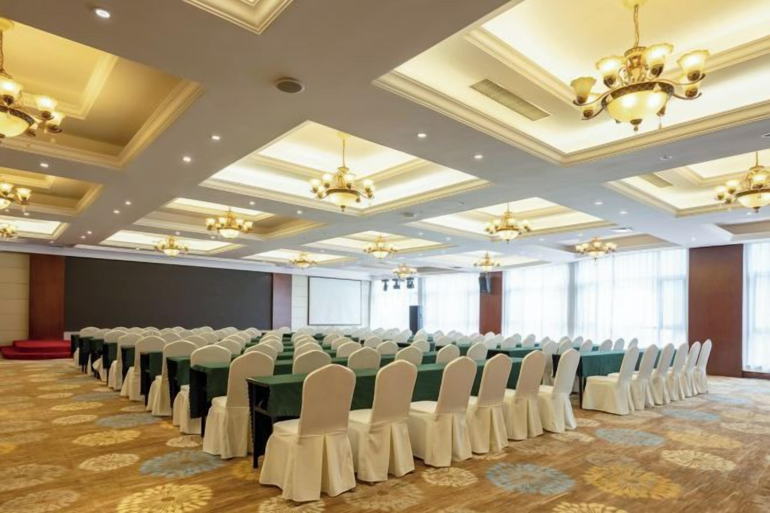 Un salón de conferencias con filas de mesas y sillas, ventanas amplias y una alfombra estampada.