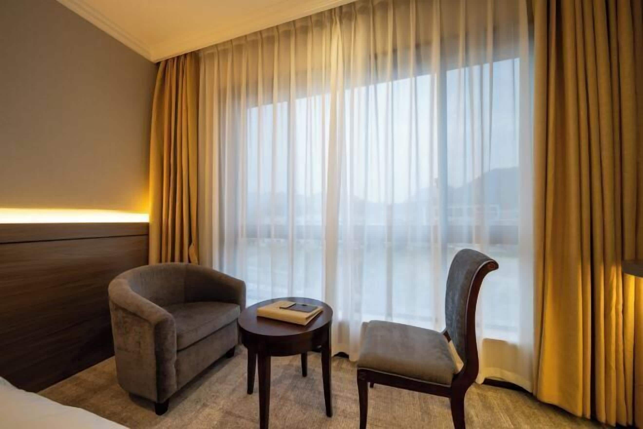 Habitación de hotel con cama, sofá, silla, una mesita pequeña y un ventanal con cortinas.