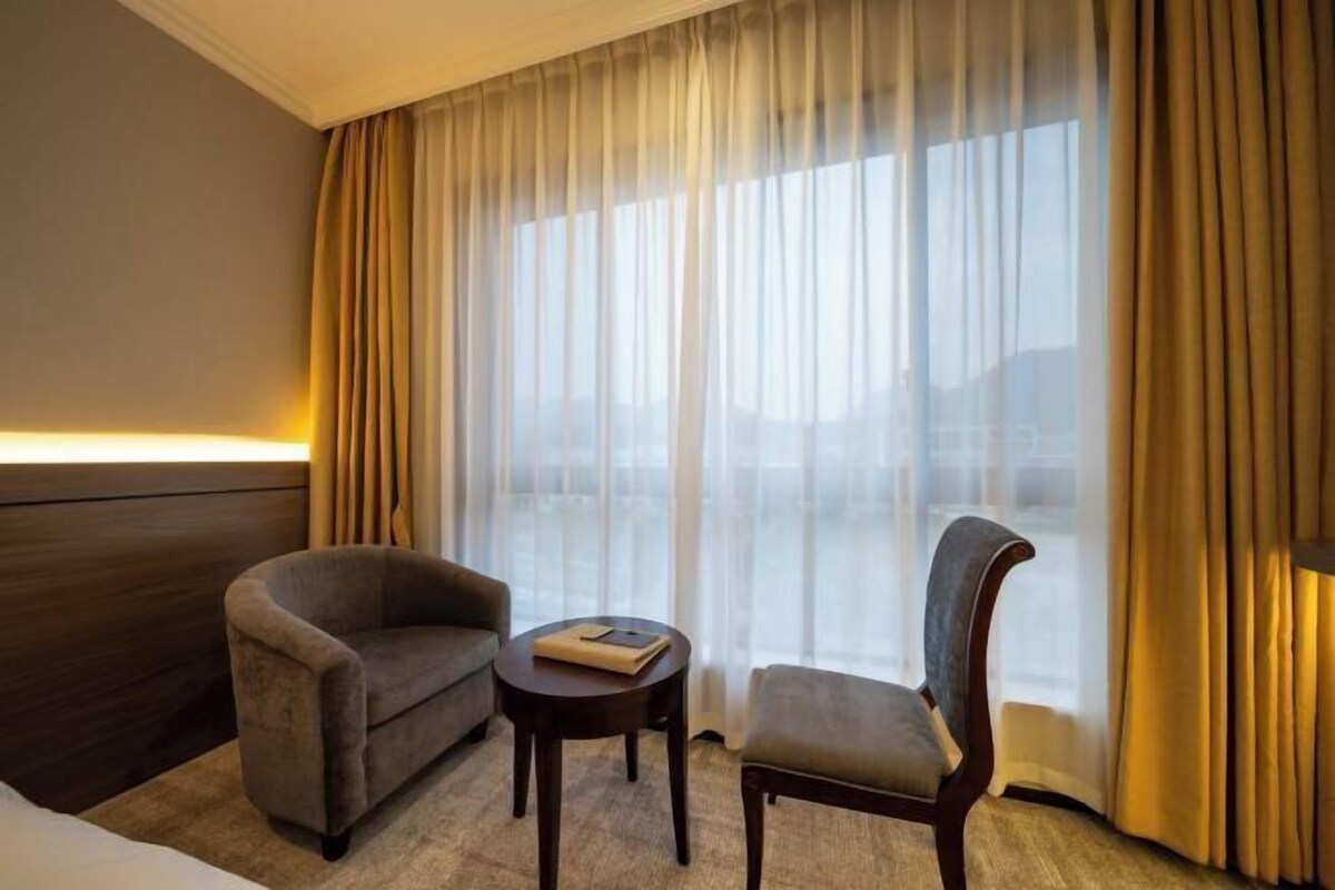 Ein Hotelzimmer mit Bett, einer Couch, einem Sessel, einem kleinen Tisch und einem großen Fenster mit Vorhängen.