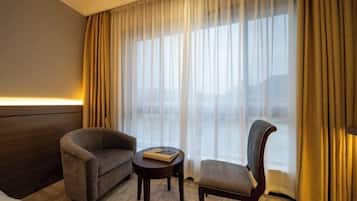 Habitación de hotel con cama, sofá, silla, una mesita pequeña y un ventanal con cortinas.