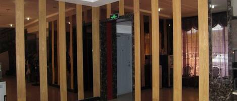 Un hall de hotel con una columna central, un mostrador de recepción y una puerta con un letrero de salida verde.