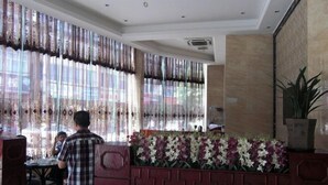 A hotel lobby with a reception desk, a seating area with a floral arrangement, and large windows with patterned curtains.