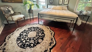 Un dormitorio con cama, una silla, una planta, un tapete y cuadros en la pared.