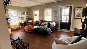 Amplia sala de estar con un sofá grande, un sillón blanco y una mesa de centro de madera. Varias ventanas dejan entrar luz natural, y una puerta da al exterior.