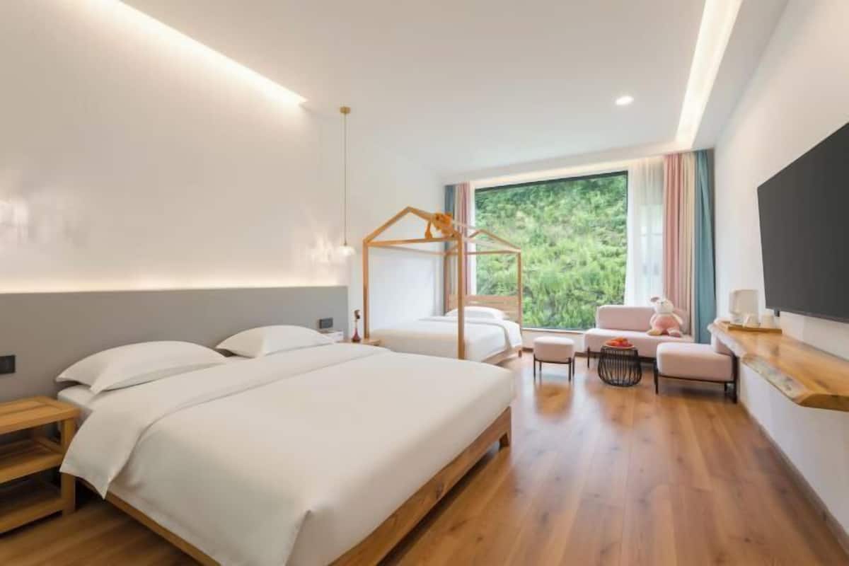 Una habitación de hotel moderna con una cama grande, un pequeño sofá, una mesita de madera y un ventanal con vistas a zonas verdes.
