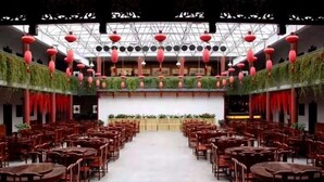Un salón de banquetes con filas de mesas y sillas de madera, faroles rojos colgando del techo y una pared verde con plantas.