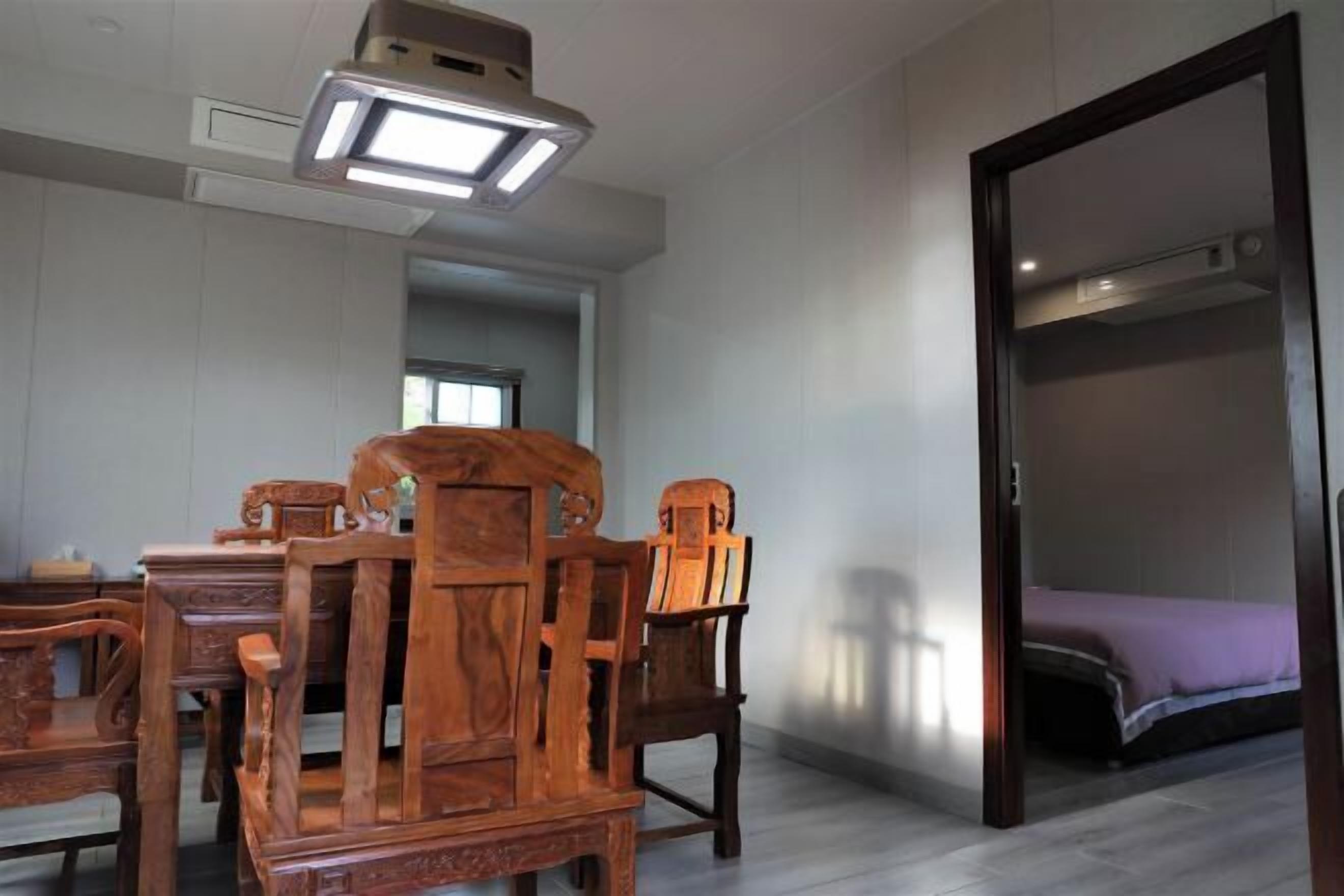 Una habitación con una mesa de madera y sillas, una cama con un edredón morado y una puerta que da a otra habitación.