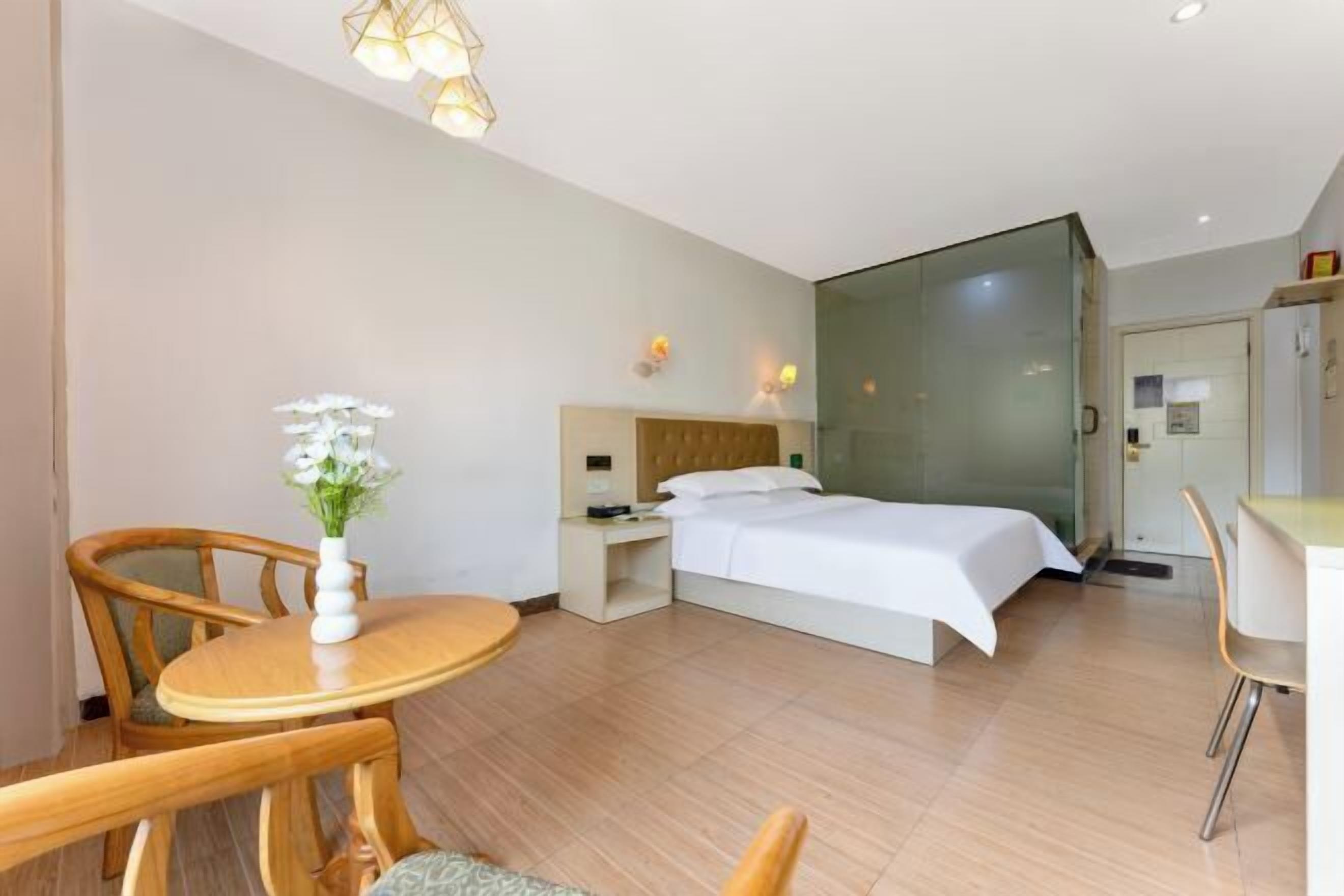 A modern hotel room with a bed, bedside table, lamp, and a dining area with chairs.
