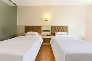 Habitación de hotel con dos camas individuales, suelo de madera, pared de color claro y mesita de noche con lámpara y teléfono.