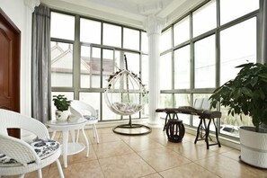 Een ruime kamer met grote ramen, een hangstoel, wit rieten meubilair en potplanten.