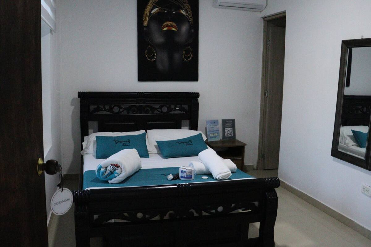 Een keurig opgemaakt bed met hoofdbord, een nachtkastje met lamp, een ingelijste foto aan de muur en een spiegel.