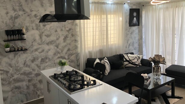 A modern living room with a black sofa, a glass coffee table, and a kitchen area with a white countertop and stove.