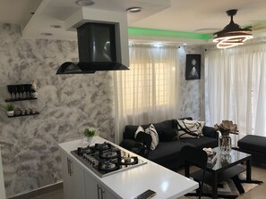 Un salon moderne avec un canapé noir, une table basse en verre et un coin cuisine doté d’un plan de travail et d’une cuisinière blancs.