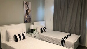A hotel room with two single beds, a nightstand with a lamp, a small potted plant, and a painting on the wall.