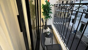 A balcony with a small table, two chairs, a potted plant, and a view of the street and buildings.