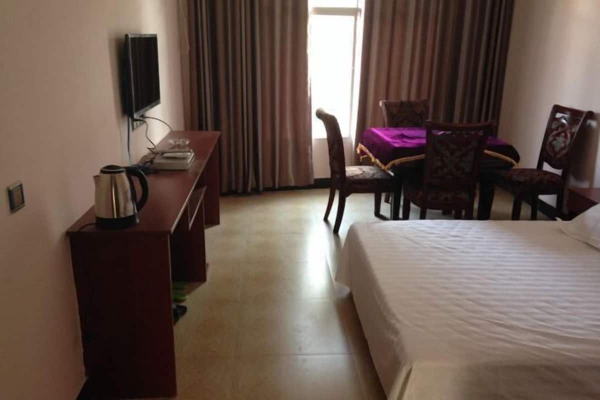 A hotel room with a bed, a desk with a kettle, a chair, a table with a purple tablecloth, and a TV mounted on the wall.