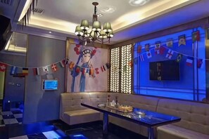 Een moderne hotellobby met een bank, een eettafel, een kroonluchter en een groot televisiescherm aan de muur.