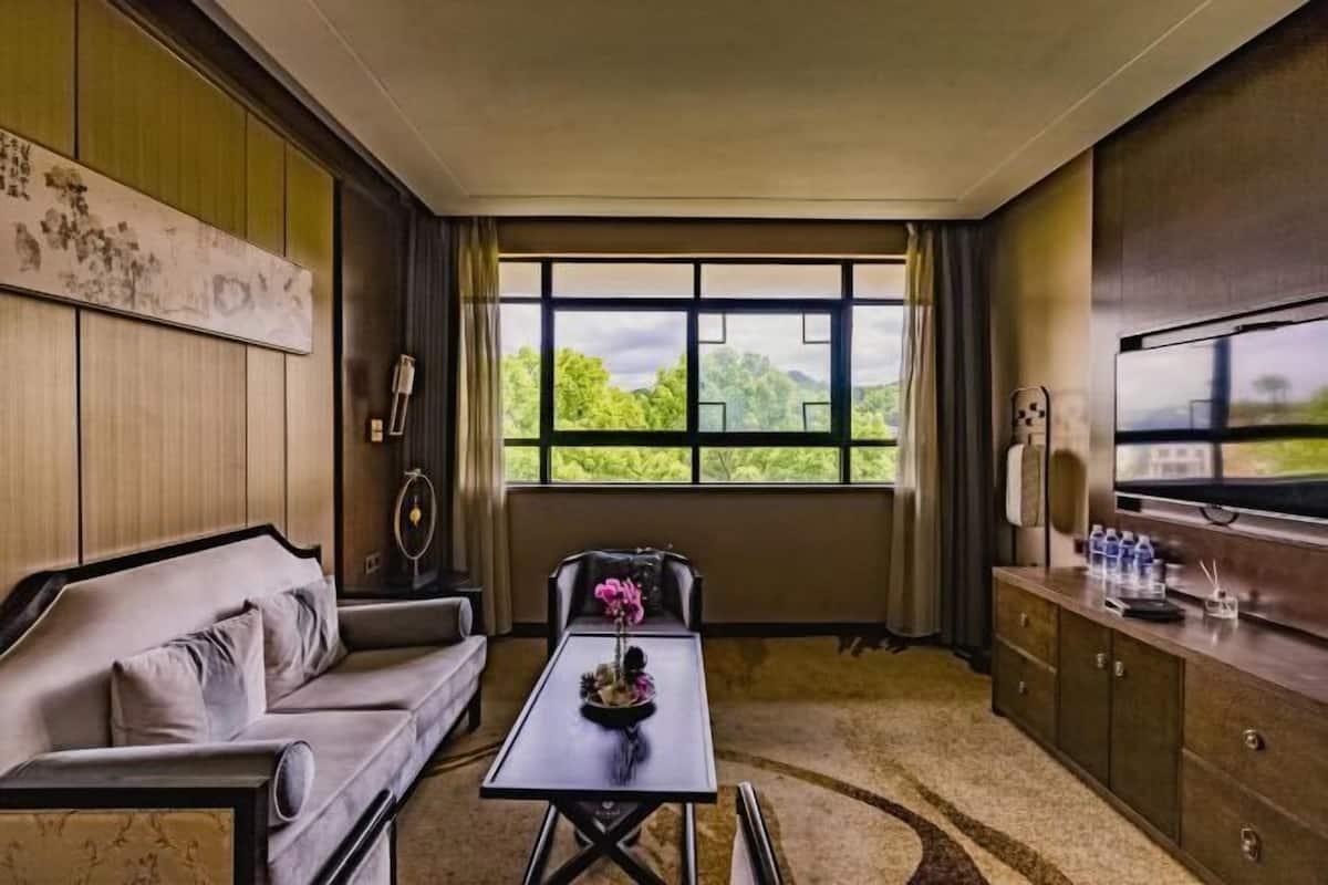 A hotel room with a sofa, a small table with a flower arrangement, a television, and a view of greenery outside.
