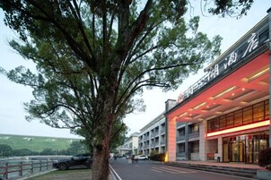 Un hotel con fachada moderna, un árbol grande en primer plano y un estacionamiento con coches.