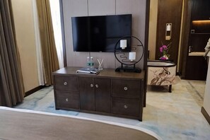 Ett hotellrum med en stor, platt-TV som är monterad på väggen, ett träbord med en fjärrkontroll, en liten prydnadsföremål och ett badrum med handfat och spegel.