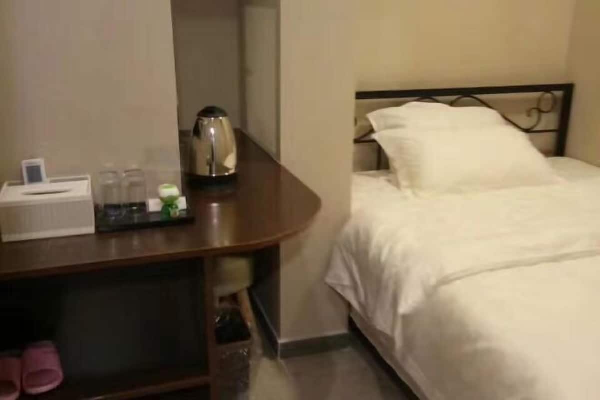 Habitación de hotel con cama, mesita de noche con lámpara, una mesita pequeña con tetera y un cesto de basura.