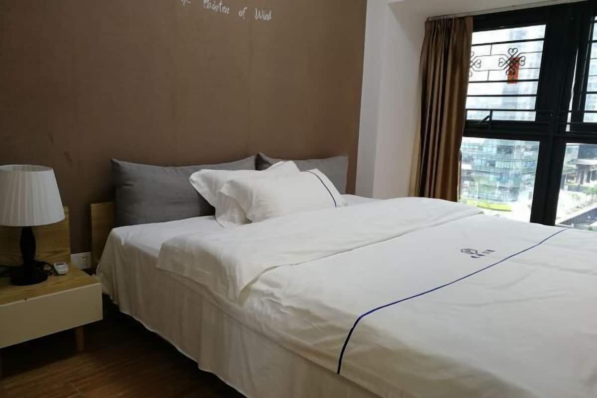 Habitación de hotel con cama, mesita de noche, lámpara y ventana con vistas a edificios.