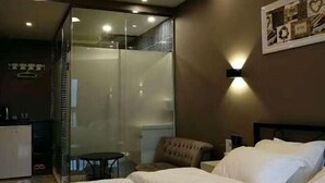 Une chambre d’hôtel comprenant un lit, une chaise, une petite table, un espace douche et un tableau accroché au mur.