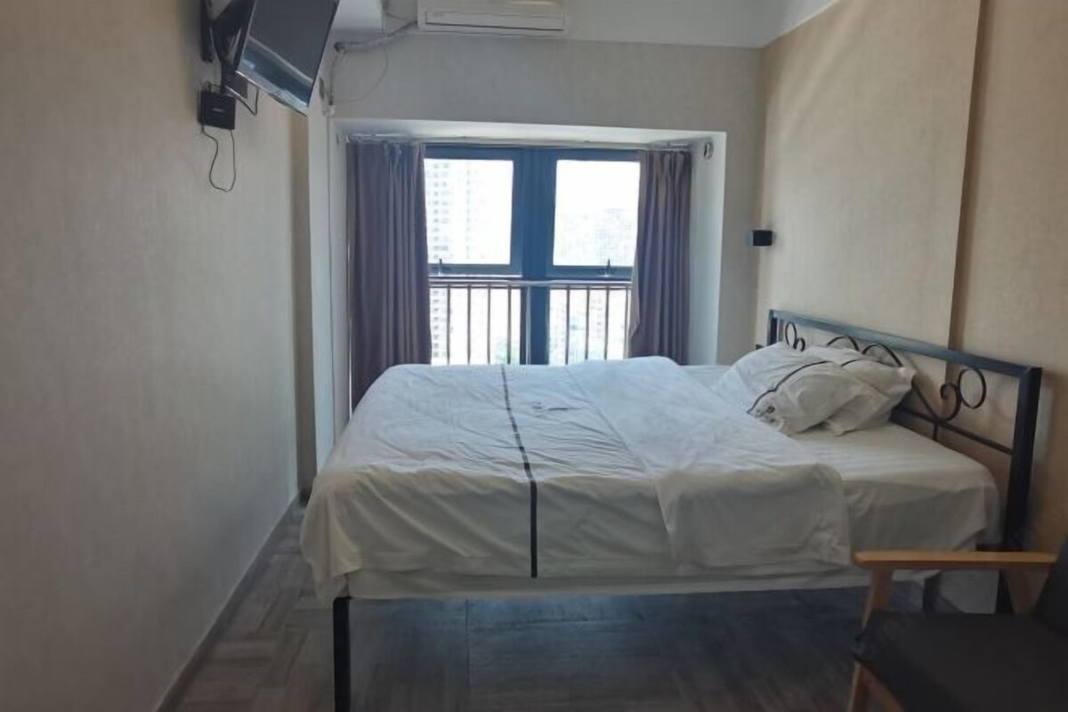 Un lit simple avec un cadre en métal, une petite table de chevet, une fenêtre avec des rideaux et un climatiseur fixé au mur.
