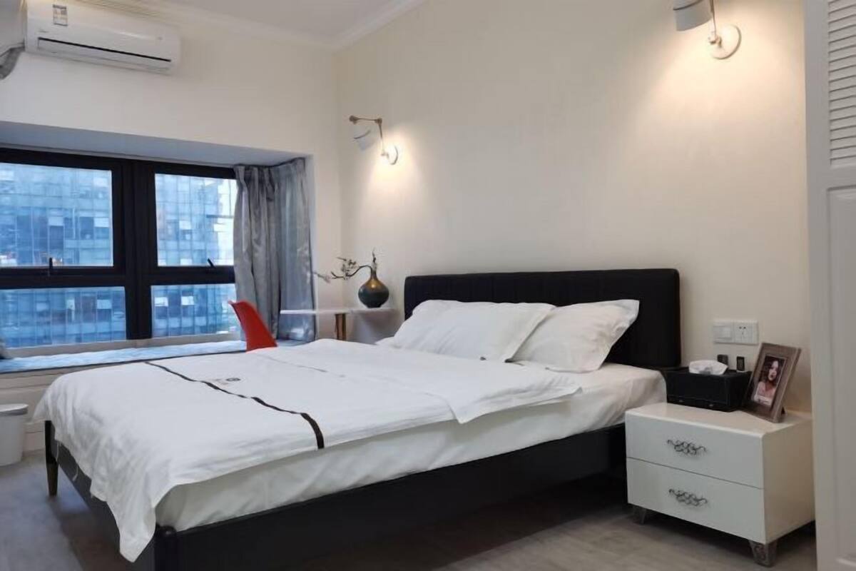 Un dormitorio moderno con una cama negra, ropa de cama blanca, una mesita de noche con lámpara y una ventana con cortinas.