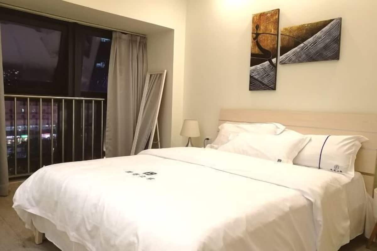 Habitación de hotel con una cama grande, una mesita de noche, una lámpara y vistas a la ciudad desde un balcón.