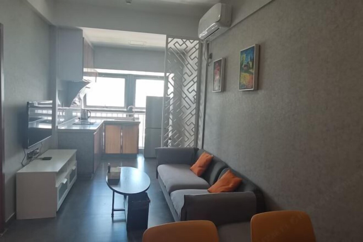 Un salon moderne avec un canapé, une table basse et un téléviseur. On aperçoit, au fond, un coin cuisine avec une fenêtre.