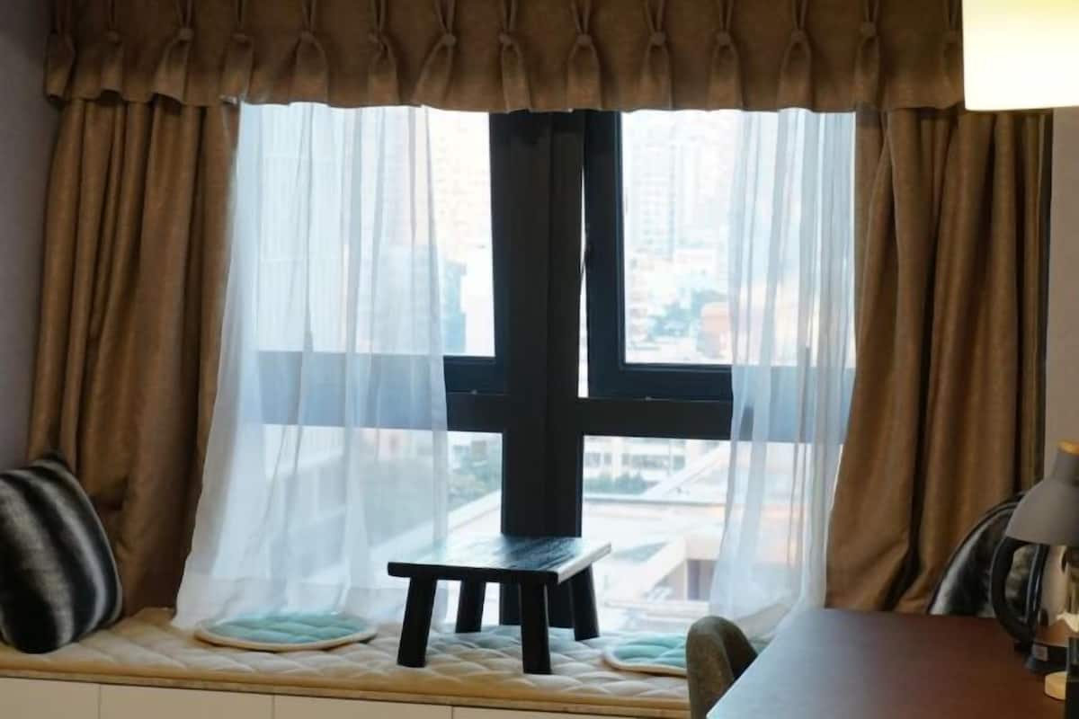 Una habitación con ventana, un taburete pequeño, un cojín y una lámpara sobre una mesa.