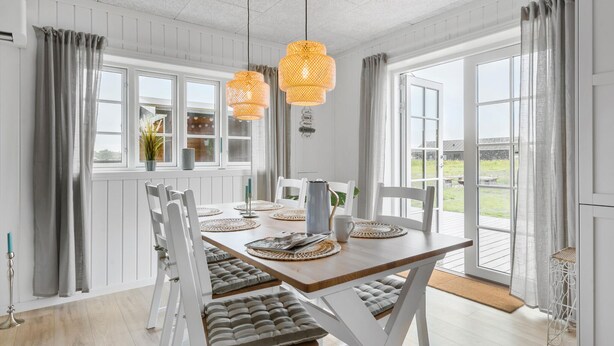 Et spiseområde med et træbord, hvide stole og en bænk. To hængende orange lamper, en hvid kande og en potteplante på vindueskarmen.