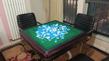 Quarto com mesa verde para jogos de tabuleiro, quatro cadeiras, janela com persianas e radiador.