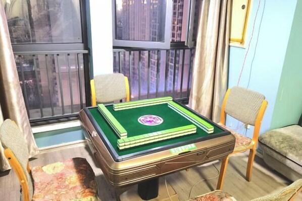 Une pièce avec une table à carreaux verts et blancs, deux chaises en bois, une fenêtre donnant sur des immeubles et un rideau.