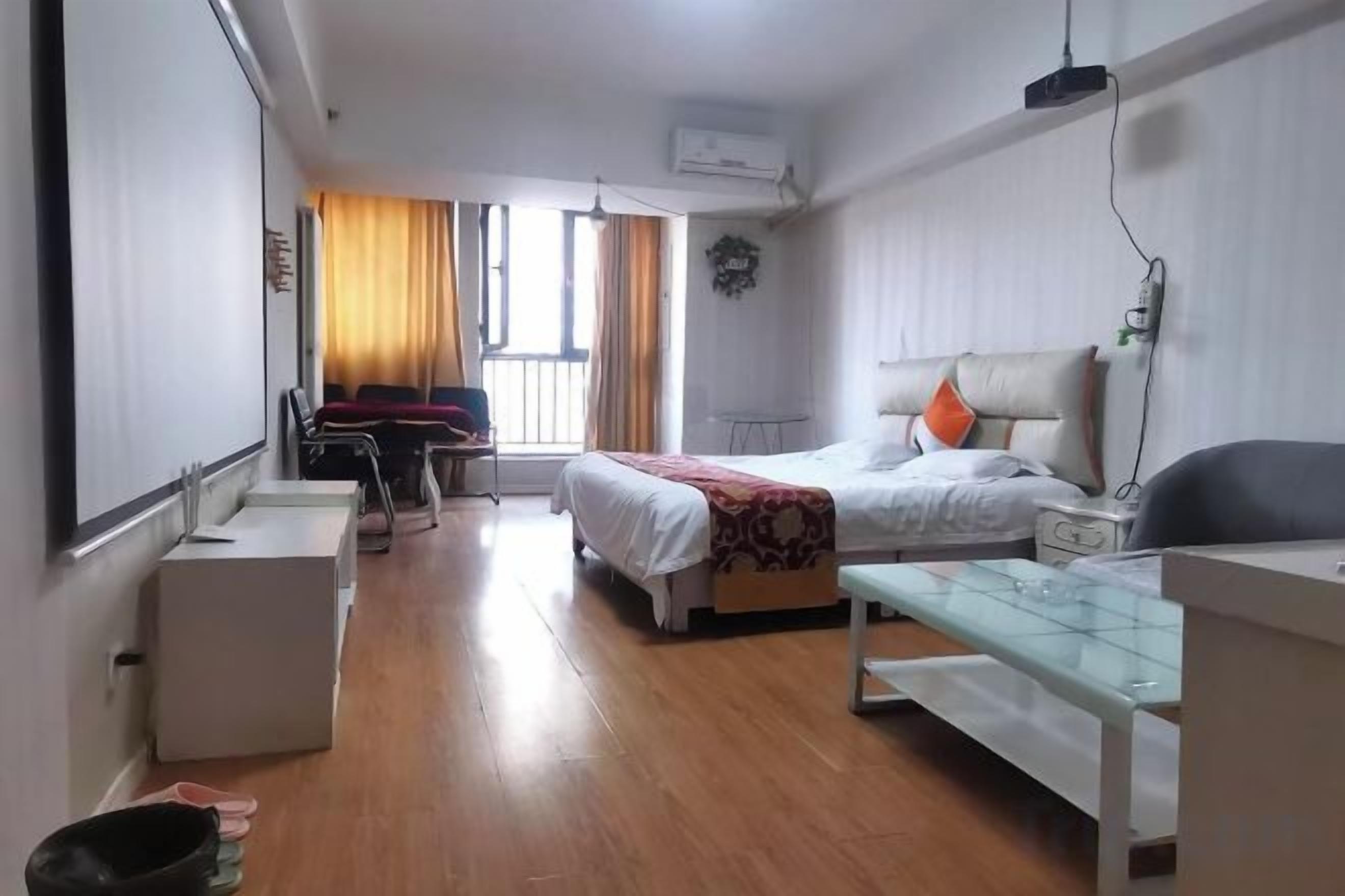 A hotel room with a bed, bedside table, a small desk, and a chair. There is a window with curtains, a wall-mounted air conditioner, and a flat-screen TV.