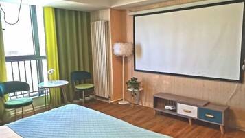 Habitación de hotel con cama, escritorio, dos sillas, una mesita, pantalla para proyector y radiador.