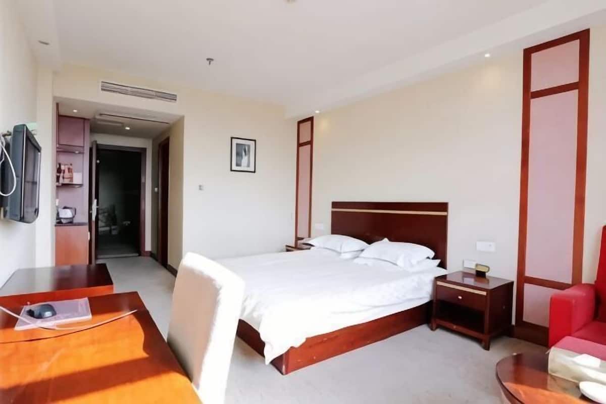 A hotel room with a wooden bed, bedside table, chair, and a desk with a chair.