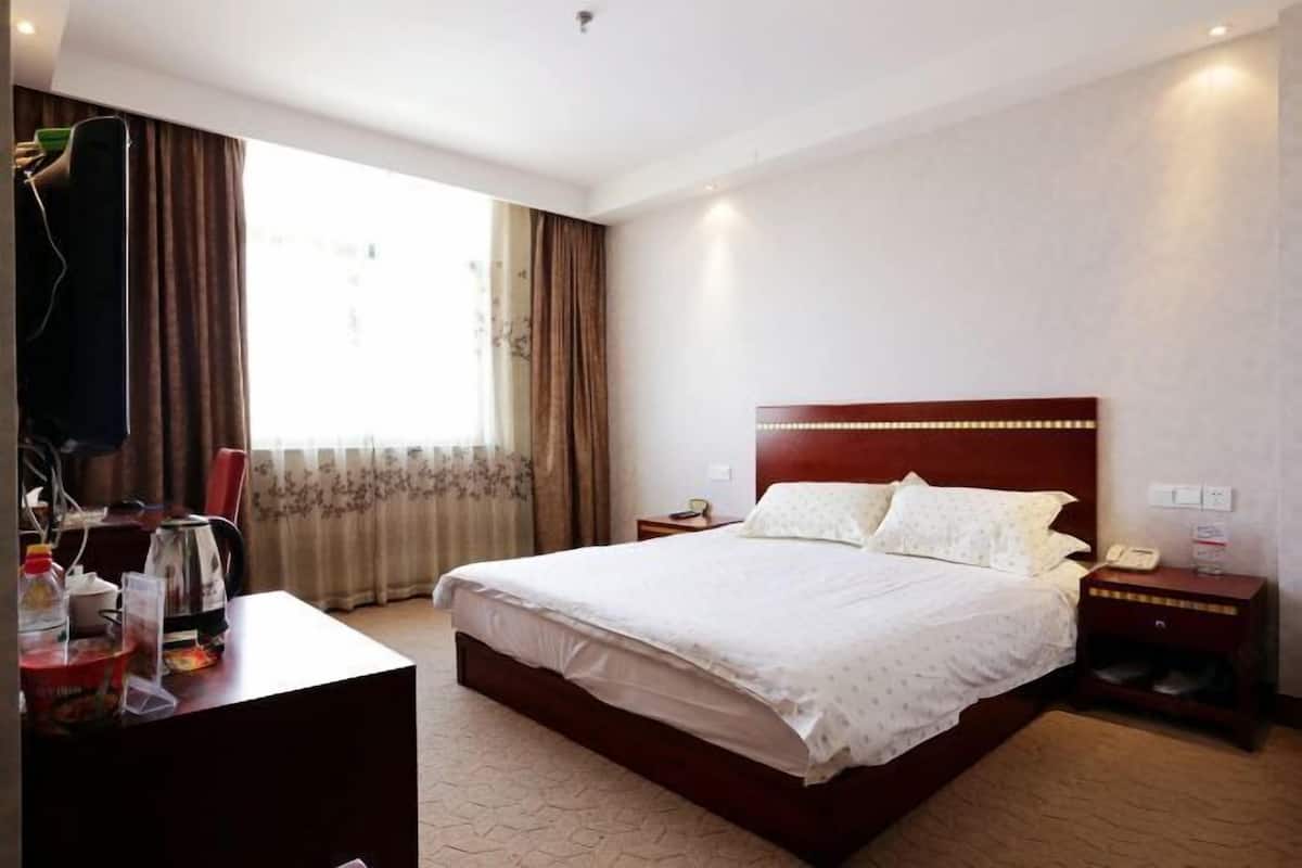 A hotel room with a large bed, a desk with a chair, a nightstand with a phone, and a window with curtains.