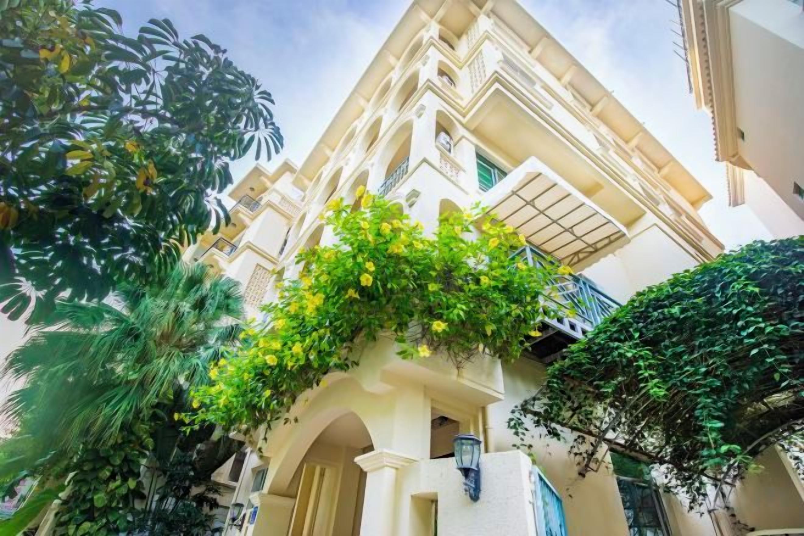 A multi-story building with balconies, arched windows, and a lush green garden area.