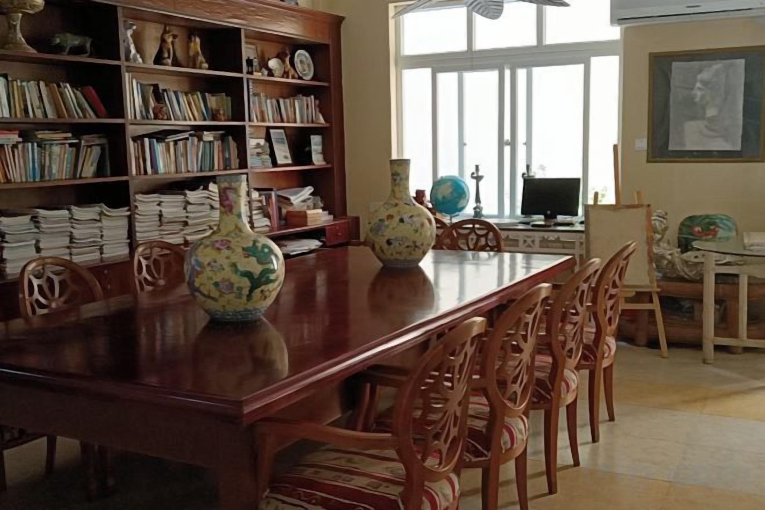 Un comedor con una mesa de madera larga, sillas, una estantería con libros y jarrones decorativos.