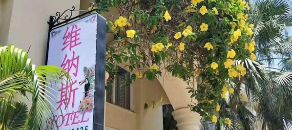 Une enseigne d’hôtel avec des caractères étrangers, un balcon orné de fleurs jaunes et des palmiers.