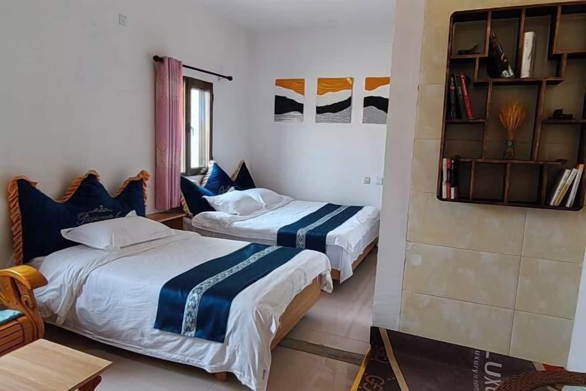A hotel room with two single beds, a wooden nightstand, a small table, a bookshelf, and a window with curtains.