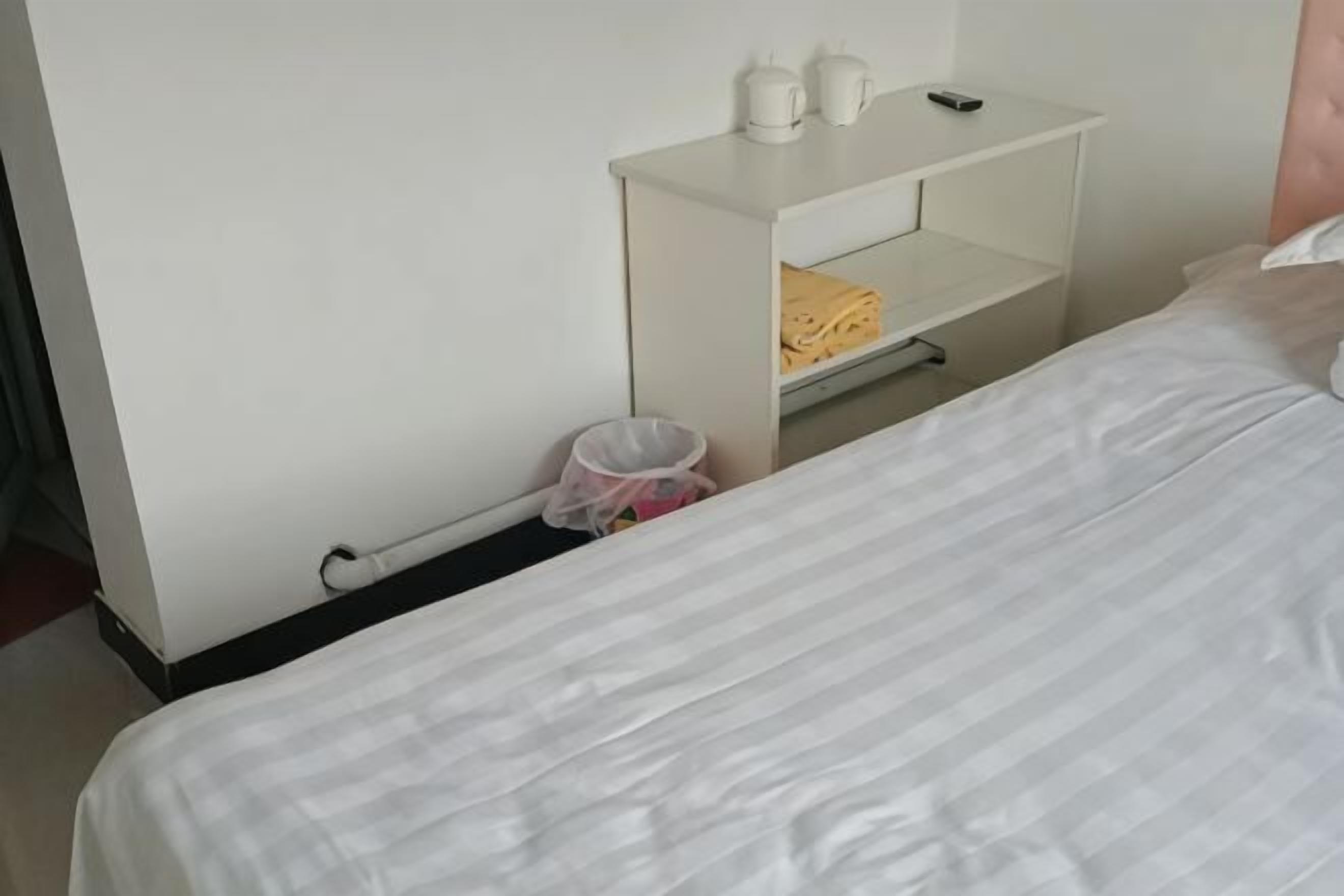 A hotel room with a bed, a white nightstand with a lamp, a shelf with folded towels, and a trash bin.