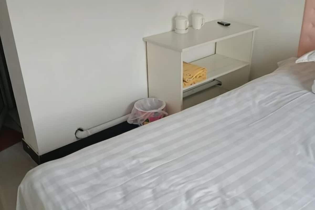 A hotel room with a bed, a white nightstand with a lamp, a shelf with folded towels, and a trash bin.