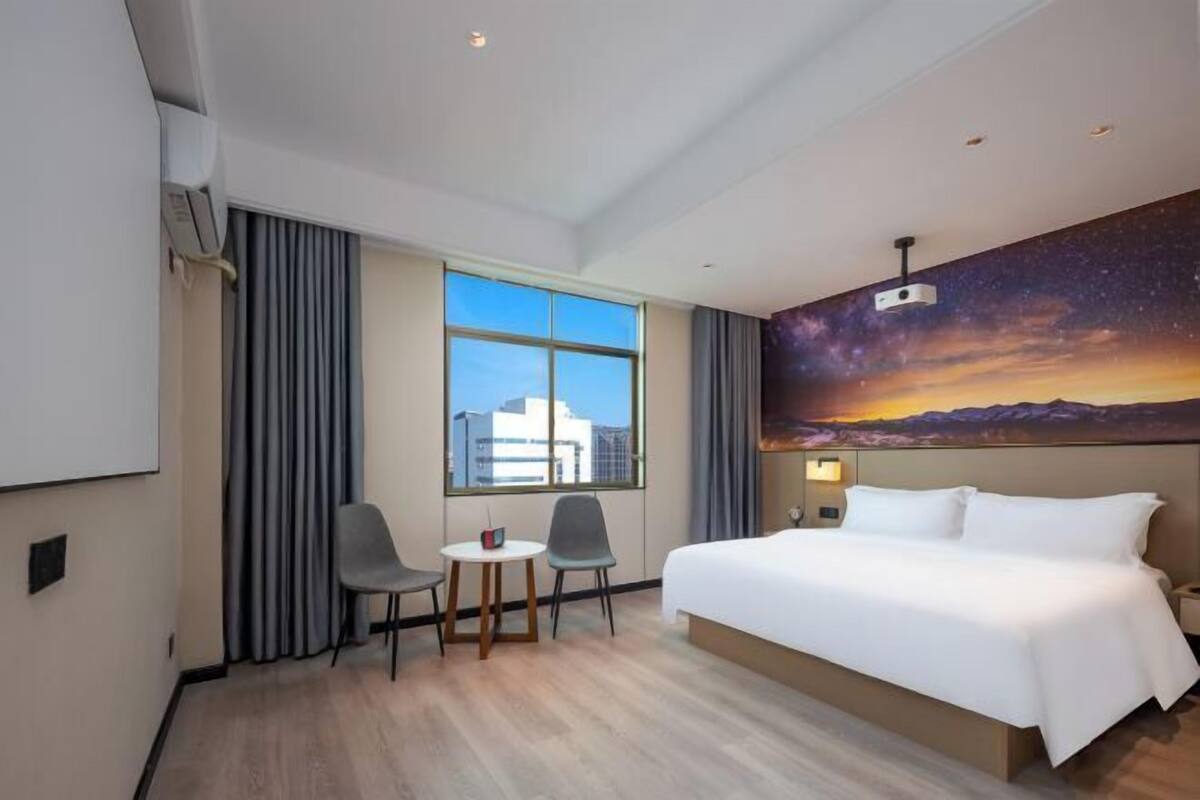 A hotel room with a large bed, a desk with two chairs, a window with curtains, and a mural of a starry sky.